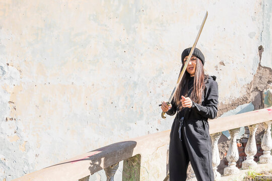 A young girl warrior in black clothes stands against the background of a concrete wall and holds a naked sword in front of her