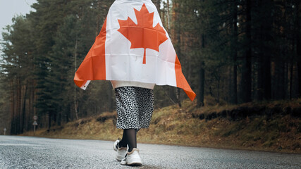 A girl in a white sweater and skirt walks through the forest holding the flag of canada