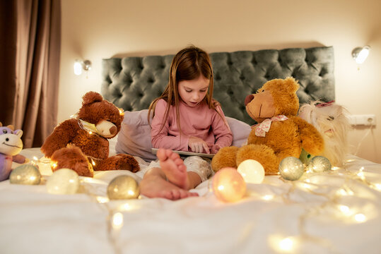 A little cute girl wearing pyjamas sitting alone on a big bed barefoot scrolling photos in her tablet having her teddybears around