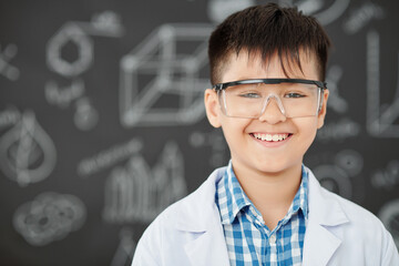 Portrait of cheerful Asian schoolboy in labcoat and plastic googles standing in chemistry class and...