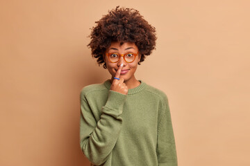 Fototapeta na wymiar Funny curly haired young woman tries to touch nose smiles happily has fun and poses in casual green wear against brown background. Pretty teenage girl in spectacles with Afro hair points to nose.