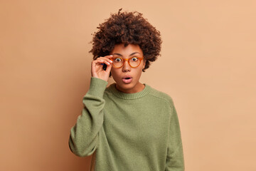Fototapeta na wymiar Emotive dark skinned woman stares at camera has eyes popped out holds breath from wonder looks scrupulously at camera wears casual sweater isolated over beige background. Human reaction concept