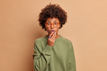 Fototapeta na wymiar Studio shot of shocked speechless curly woman keeps hand near mouth stands impressed indoor stares through spectacles wears casual jumper isolated over beige background hears unbelievable news