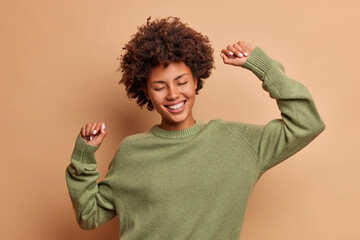 Horizontal shot of happy curly haired Afro American woman feels lively and dances carefree keeps...