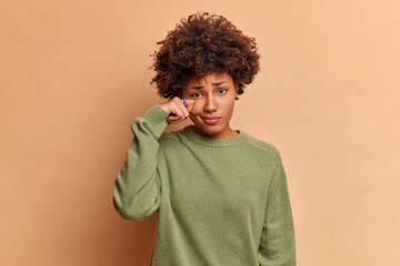 Fototapeta na wymiar Gloomy dissatisfied curly haired woman wipes teardrops under eye expresses sadness and depression feels unhappy wears casual jumper isolated over brown studio wall. Negative emotions concept