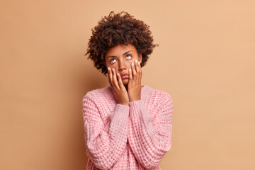 Fototapeta na wymiar Upset bored Afro American woman keeps hands on cheeks suffers from boredom doesnt know what to do has dull expression stays at home alone dressed in casual sweater isolated over beige background