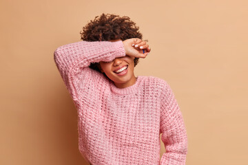 Overjoyed young woman laughs happily and smiles broadly hides eyes with arm being in good mood hears hilarious story laughs out as hears anecdote dressed in jumper isolated over brown background