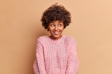Obraz na płótnie Canvas Photo of glad millennial dark skinned woman with Afro hair smiles gently and looks away being in high spirit expresses positive emotions isolated over beige studio background. Happiness concept