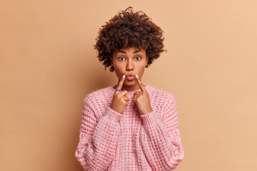 Fototapeta na wymiar Portrait of lovely woman keeps fingers near folded lips has direct gaze at camera dressed in loose knitted sweater poses against brown background forces smile. Human face expressions concept