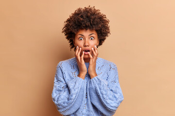 Impressed dark skinned woman with curly hair grabs face as sees something shocking and breathtaking popping eyes at camera hears horrible news dressed in casual jumper poses against brown wall