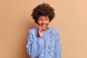 Fototapeta na wymiar Dreamy thoughtful woman with Afro hair looks away smiles gently concentrated aside grins positively dressed in blue jumper poses against brown background. Pleasant thoughts and feelings concept