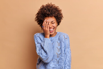 Happy carefree dark skinned woman with curly hair laughs out loudly from funny joke makes face palm watches something hilarious wears casual jumper isolated over beige background. Sincere emotions