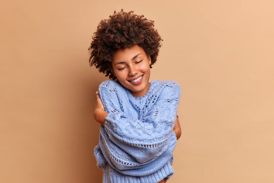 Pleased smiling woman with bushy curly hair embraces herself with love enjoys softness of new jumper feels comfortable and delighted closes eyes in satisfaction isolated over beige background.