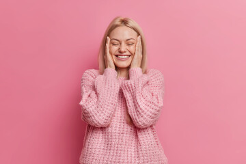 Positive sincere blonde woman keeps hands on cheeks closes eyes with pleasure dressed in casual winter jumper grins from happiness isolated over rosy studio wall. People and emotions concept