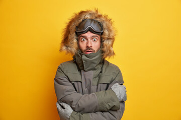 Winter vacation and extreme sport concept. Shocked snowboarder trembles from cold embraces himself as tries to warm during frosty day has mountain ski resort isolated over yellow studio wall