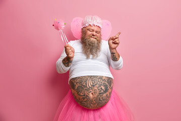 Joyful bearded adult man fairy dances carefree has fun on costume party holds magic wand closes eyes shows tattooed belly poses against rosy background. People holiday and entertainment concept. - obrazy, fototapety, plakaty