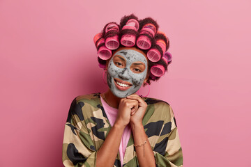 Good looking smiling housewife undergoes beauty treatments applies face mask and makes hairdo for special occasion keeps hands together near face dressed in casual wear isolated over pink background