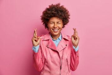 Superstitious African American woman hopes dreams come true smiles positively dressed in stylish jacket has beliefs in better isolated over rosy background waits for miracle. Body language concept