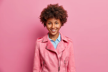 Obraz na płótnie Canvas Stylish pretty dark skinned woman with curly hair pleasant smile dressed in fashionable rosy jacket expresses positive emotions shows optimism poses indoor. Happy emotions and feelings concept