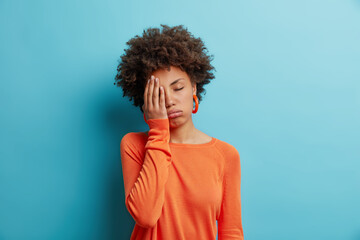 Fototapeta na wymiar Exhausted unhappy woman makes face palm and sighs from tiredness has sleepy expression fed up of working without rest wears orange jumper in one color with earrings. Upset depressed female model