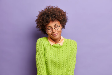 Dreamy satisfied young Afro American woman stands with closed eyes smiles gently recalls something pleasant tilts head wears round spectacles and casual jumper isolated over purple background