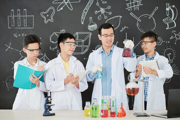 Smiling young Asian chemistry teacher showing what liquids he is going to mix to show chemical...
