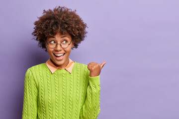 Happy good looking ethnic woman with Afro hair points away on blank space shows place for your advertising content or commercial text dressed in casual jumper isolated over purple backround. - obrazy, fototapety, plakaty