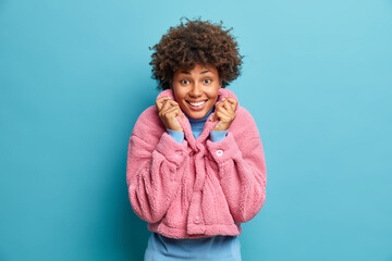 Portrait of curly haired woman dressed in pink eco coat smiles pleasantly at camera enjoys good day isolated over blue background. Optimistic stylish ethnic lady happy to buy new winter clothes