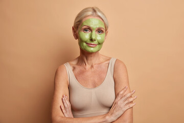 Confident charming woman with blonde hair keeps arms crossed wears minimal makeup applies green face mask dressed in casual top isolated over brown background. Beauty aging perfect skin concept