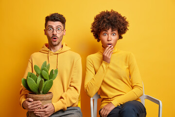 Handsome shocked man and Afro American woman sit next to each other on chairs hear unbelievable news dressed casually isolated over yellow background afraid of mistake. Birthday holiday concept