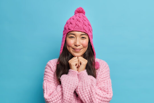 Adorable brunette Asian girl keeps hands under chin looks pleasantly at camera wears winter hat and sweater ready for walk during cold day isolated over blue background. Season and fashion concept