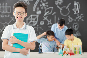 Portrait of happy Vietnamese school student in glasses holding notebook and smiling at camera when standing in science class