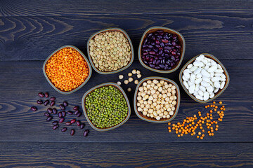 Assortment of colorful legumes in bowls: lentils, beans, mung and chickpeas on a dark rustic background, top view.  Healthy vegan protein food.