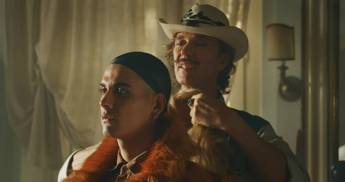 Cinematic close up shot of carefree happy smiling homosexual male gay is helping to put on a wig to his transgender woman partner and giving affective kiss as sign of forever love at home.