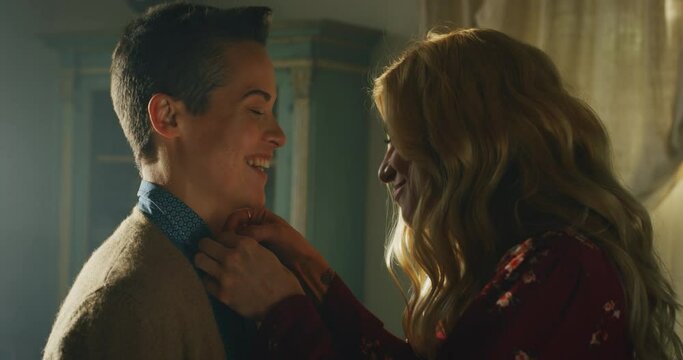 Cinematic close up shot of young carefree happy smiling homosexual blond hair female gay is helping buttoning a shirt to her transgender man and giving affective kiss as sign of forever love at home.