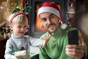 Christmas online video chatting, online greetings. Cheerful father and son using smartphone for virtual call and congratulations family and friends or making selfie photo. New year home celebration.