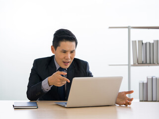 Young asian businessman in black suit frowning and pointing finger at a computer monitor. Concept of teleconferencing through wireless communication.