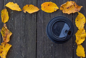 A black paper Cup of coffee stands on a wooden table on the street around an ornament of yellow leaves taken from above