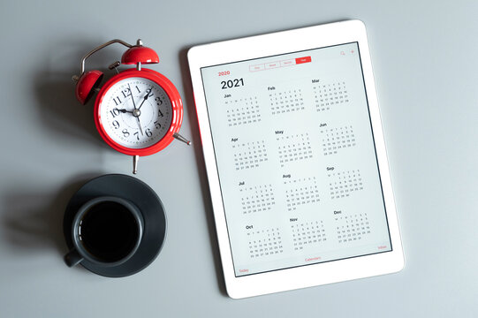 a tablet with an open calendar for 2021 year, a cup of coffee,  and a red alarm clock on a gray background