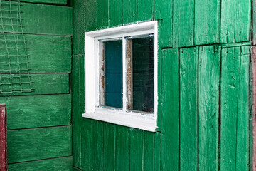 Obraz na płótnie Canvas An old wooden small window with a frame of boards painted white and thin glass. On a battered wall of wooden planks with green paint. The wall of a dilapidated building with a rough surface