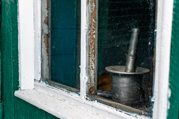 An old wooden small window with a frame of boards painted white and thin glass. On a battered wall of wooden planks with green paint. The wall of a dilapidated building with a rough surface