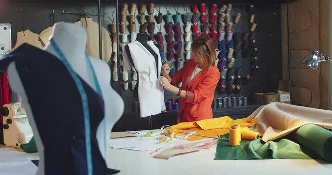 Busy young female fashion designer working in design studio with clothes on mannequin and taking measurements on fabric. Caucasian beautiful woman couturier working in atelier. Business concept