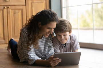 Smiling young Caucasian mother and small 7s son lying on floor at home have fun using modern tablet gadget together. Happy mom and little boy child look at pad screen watch video online or play game.
