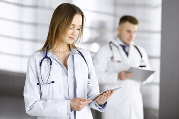 Professional woman doctor with a stethoscope is using a computer tablet, while she is standing together with her colleague in a clinic. Young doctors at work in a hospital. Medicine and healthcare