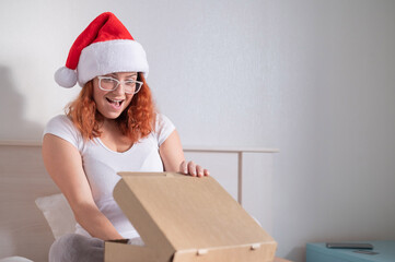 An excited red-haired woman in a Santa Claus hat sits in bed and opening a box with an order. Online shopping concept
