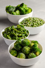 A mix of frozen green vegetables: beans peas broccoli Brussels sprouts in bowls on a grey back