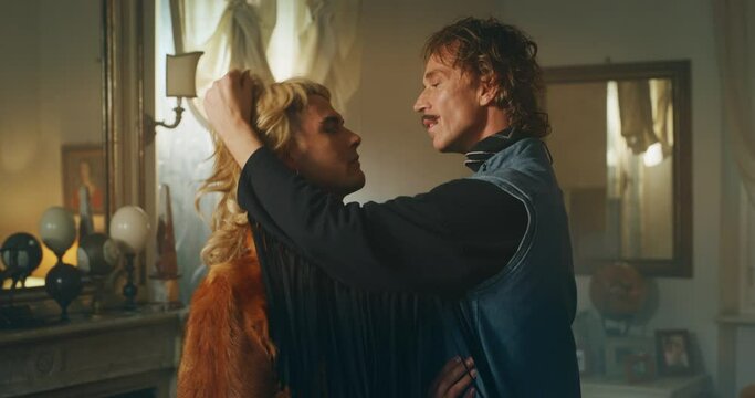 Cinematic shot of carefree happy smiling homosexual male gay dressed as cowboy is taking off a wig of his transgender woman partner and giving passion kiss as sign of forever love at home.