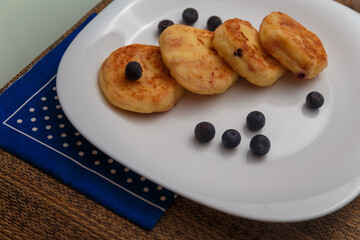 Traditional syrniki or cottage cheese pancakes with blueberries on white plate with blue napkin on kitchen table, close up.