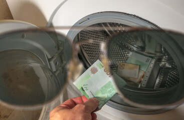 Money laundering concept, a man hand adding the 100 euro banknote into a pile of euro banknotes to be cleaned inside the washing machine, scene is seen through black criminal glasses.