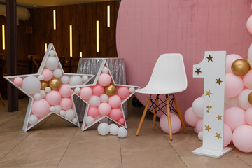 Beautiful festive decorations, pink and grey balloons, wooden stars, white chair and number one on wooden round background. Little 1 year old girl birthday party photo zone.